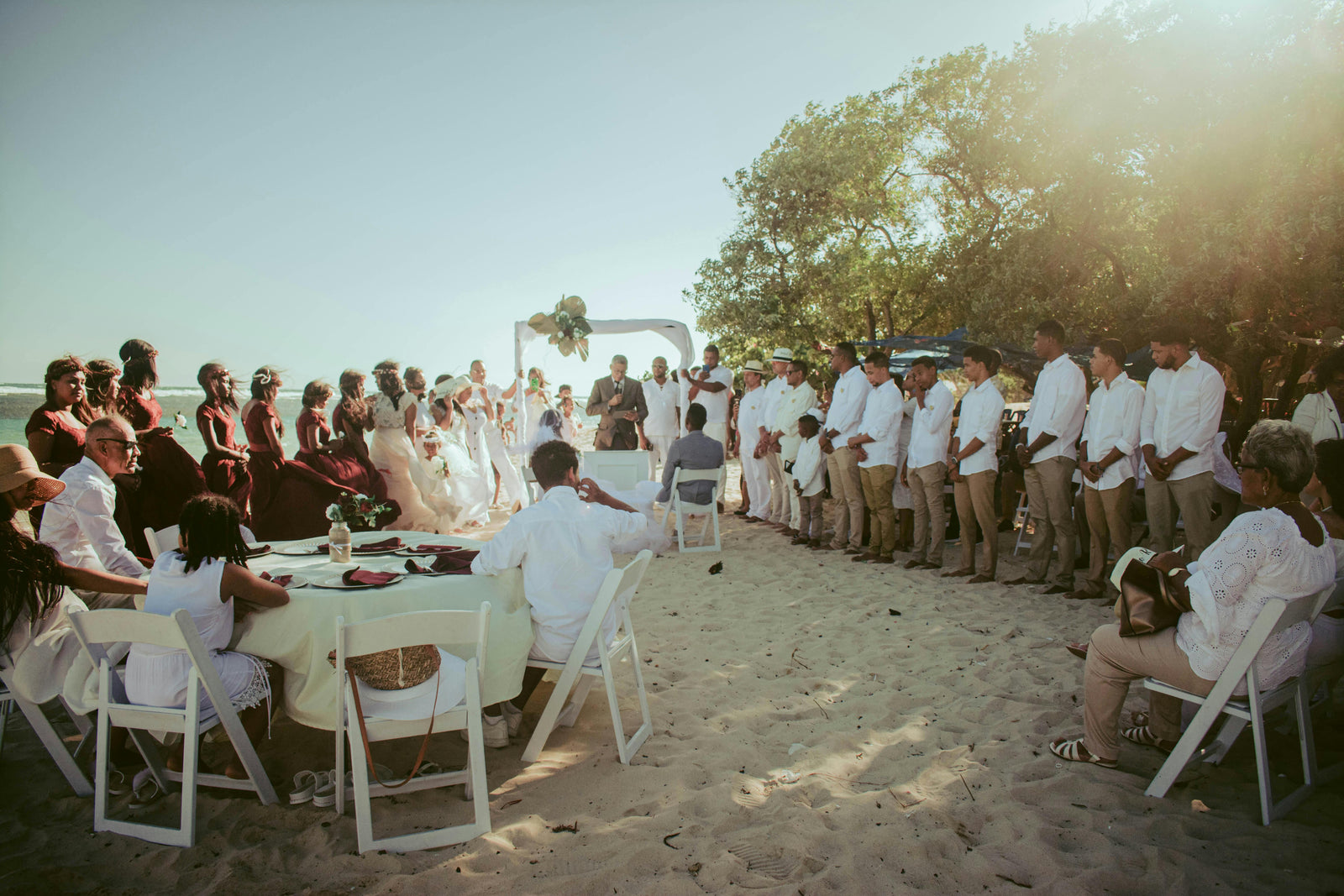 Group of people gathered for a wedding on a sandy beach with trees in the background.