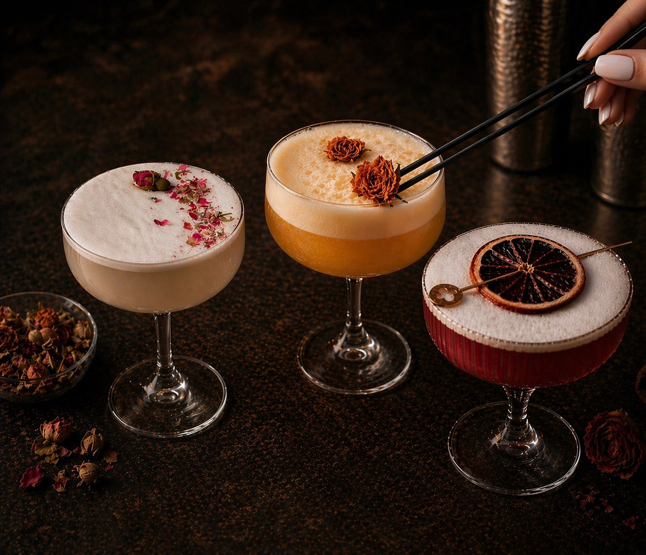 Three cocktails with decorative flower and dried fruit garnishes on a dark surface