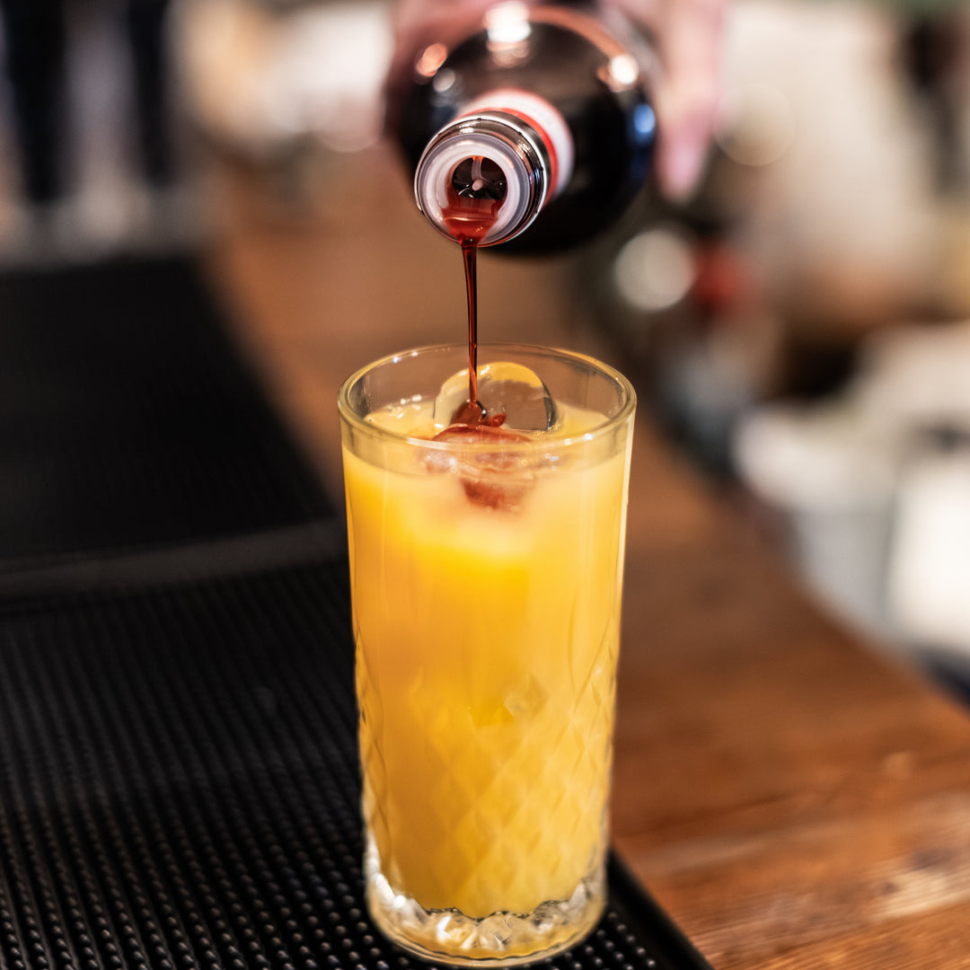 Person pouring cocktail syrup into a glass of yellow fruit juice on a bar.