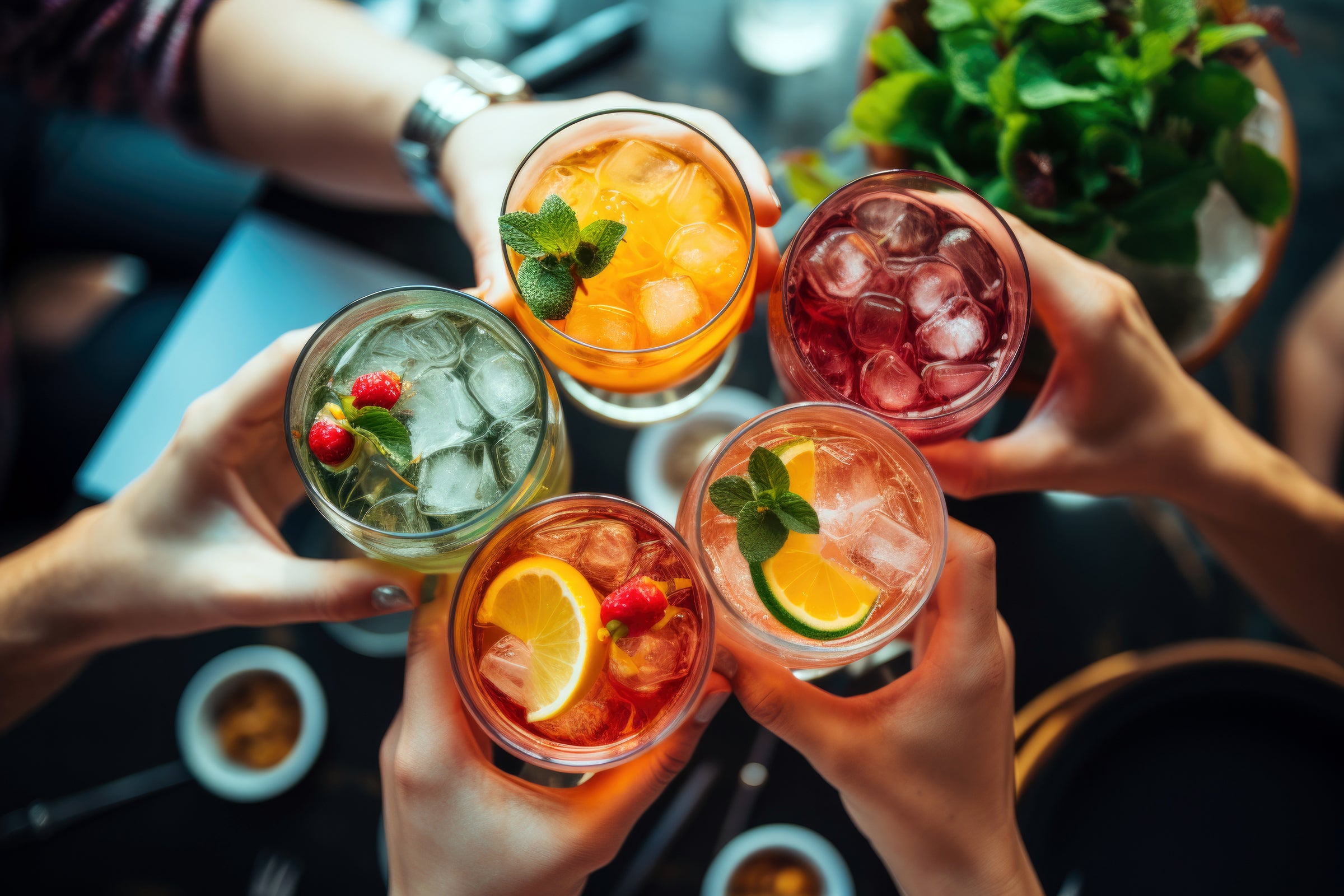 Five hands holding glasses of colorful cocktails with ice and garnishes.