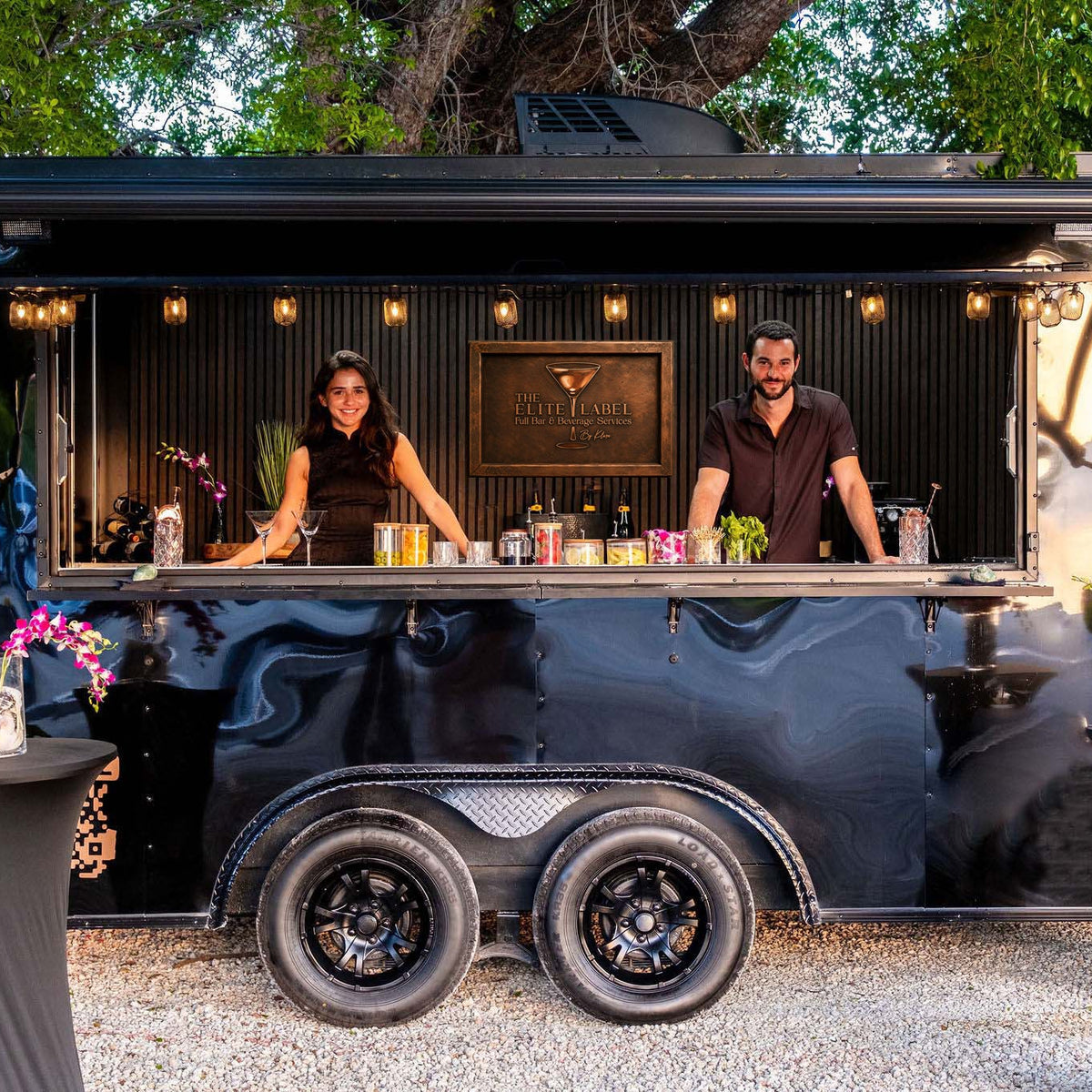 Two bartenders standing behind a mobile bar trailer with plants and garnishes around.