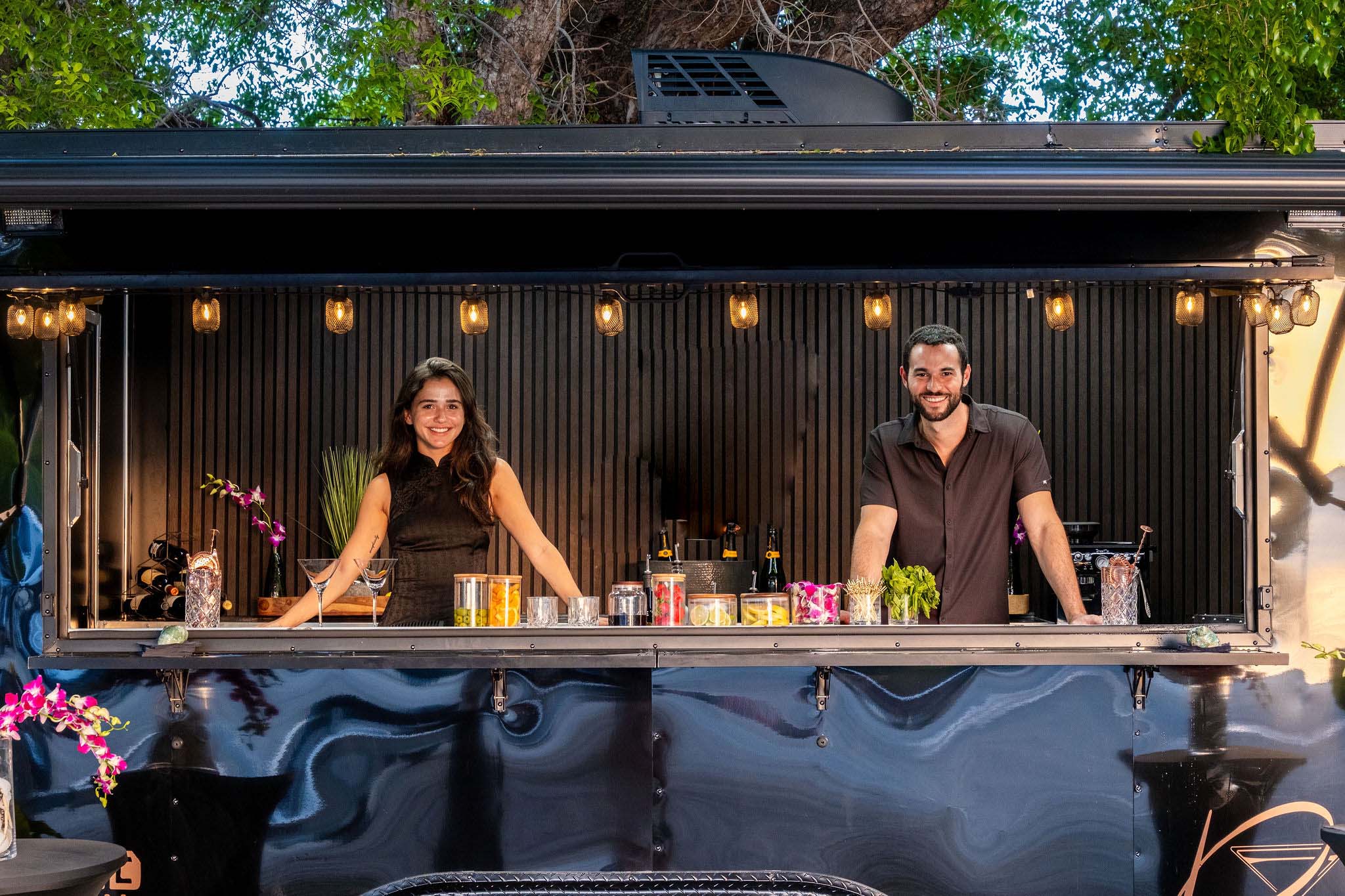 Two bartenders in a mobile bar trailer with drinks and decor in an outdoor setting.