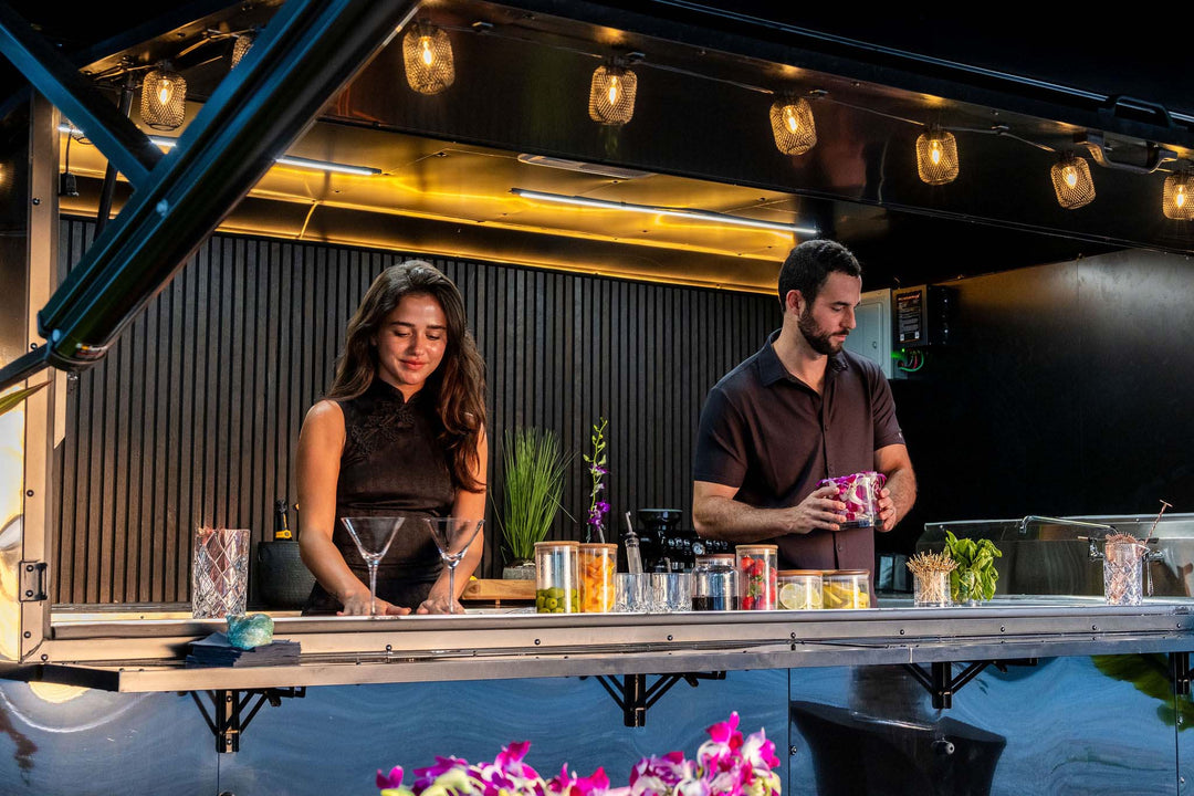 Two bartenders behind a Mobile bar counter with drinks and decor.