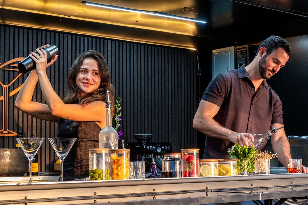 Two bartenders preparing drinks behind a mobile bar counter with various ingredients and glasses.