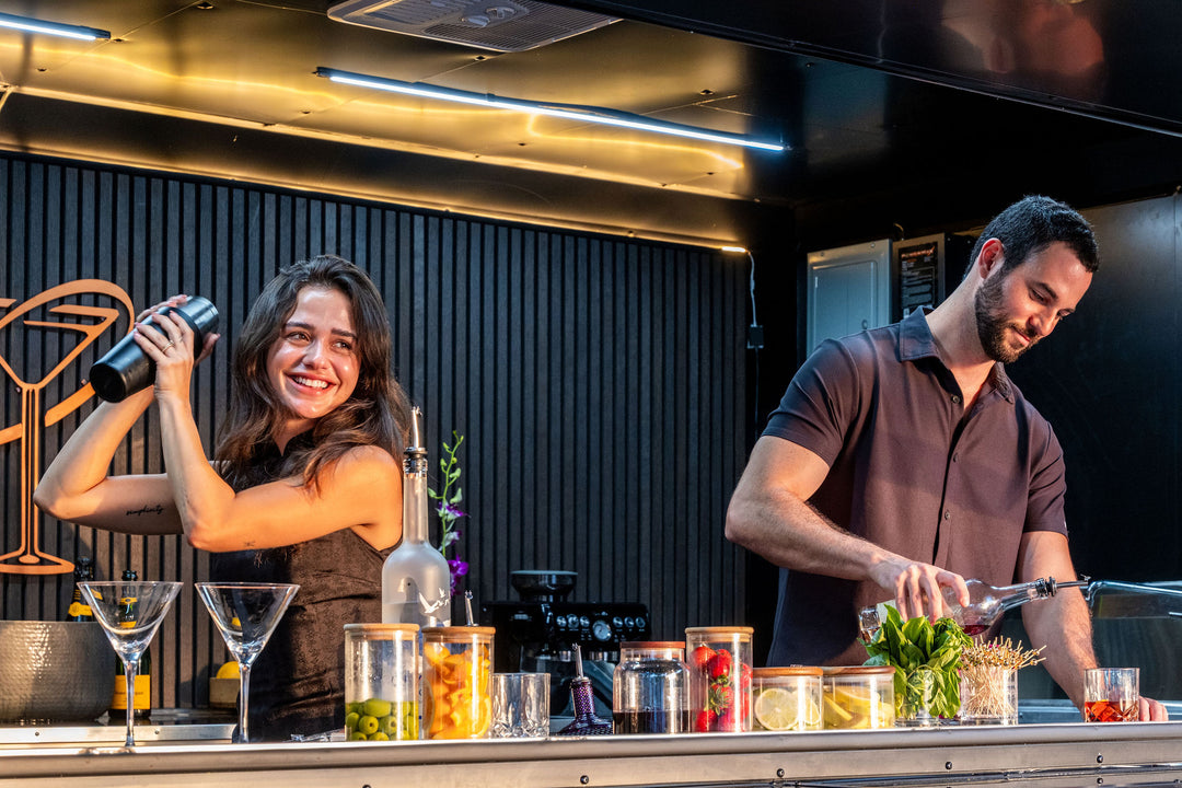 Two bartenders preparing drinks at a mobile bar counter with various ingredients and glasses.