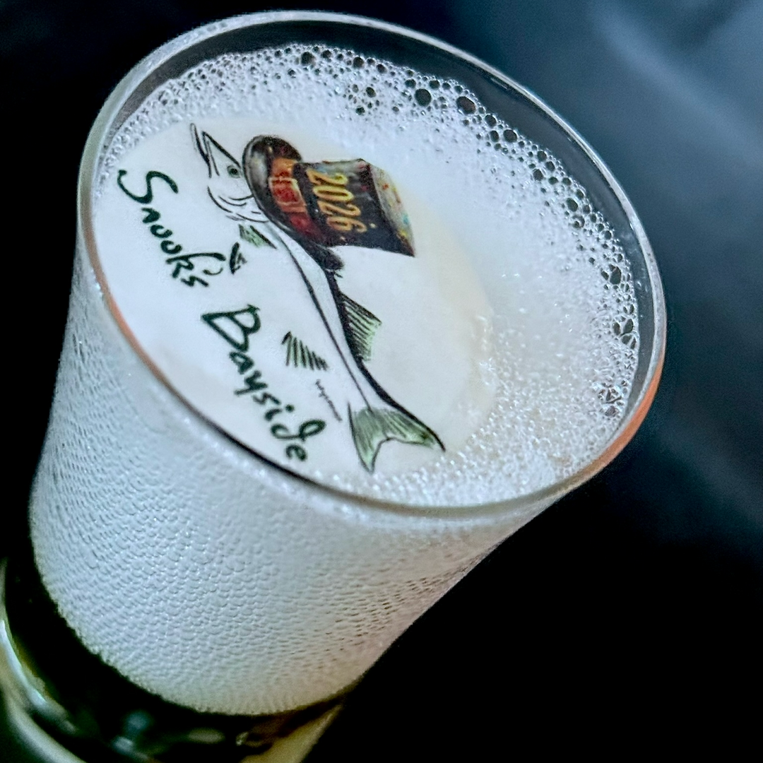 Close-up of a glass of beer with foam, featuring a branded edible cocktail topper label on a dark background.
