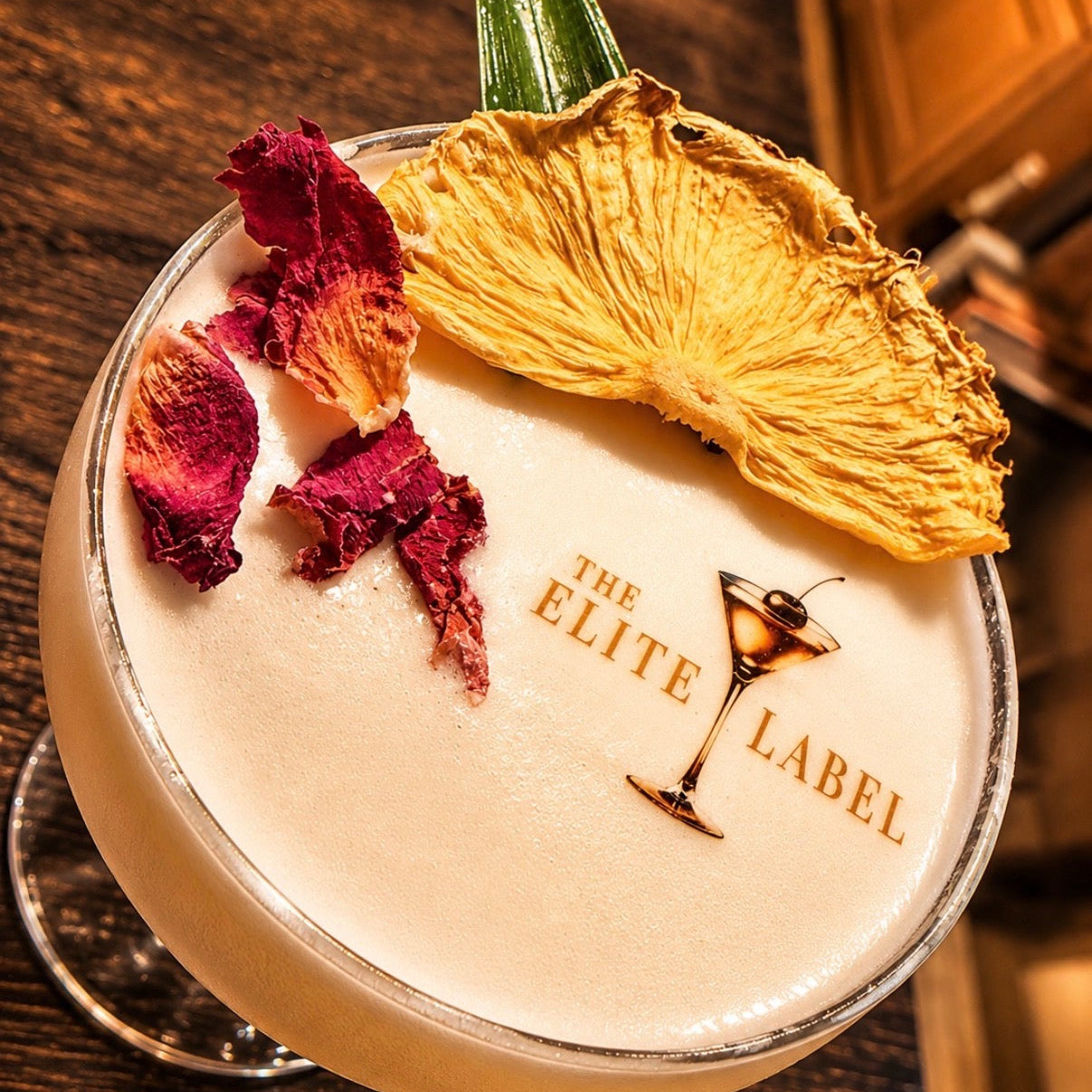 Cocktail with dehydrate pinapple garnish and floral botanicals and 'The Elite Label' drink print branding on a wooden surface.