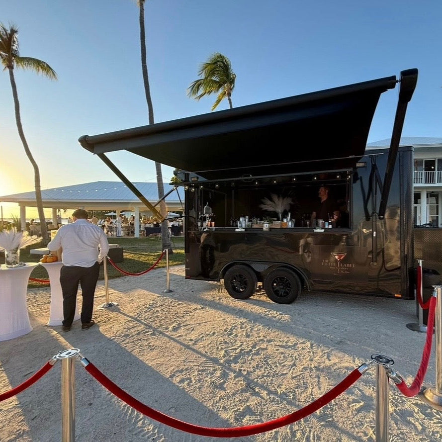 Mobile bar trailer on a sandy area with palm trees and a building in the background