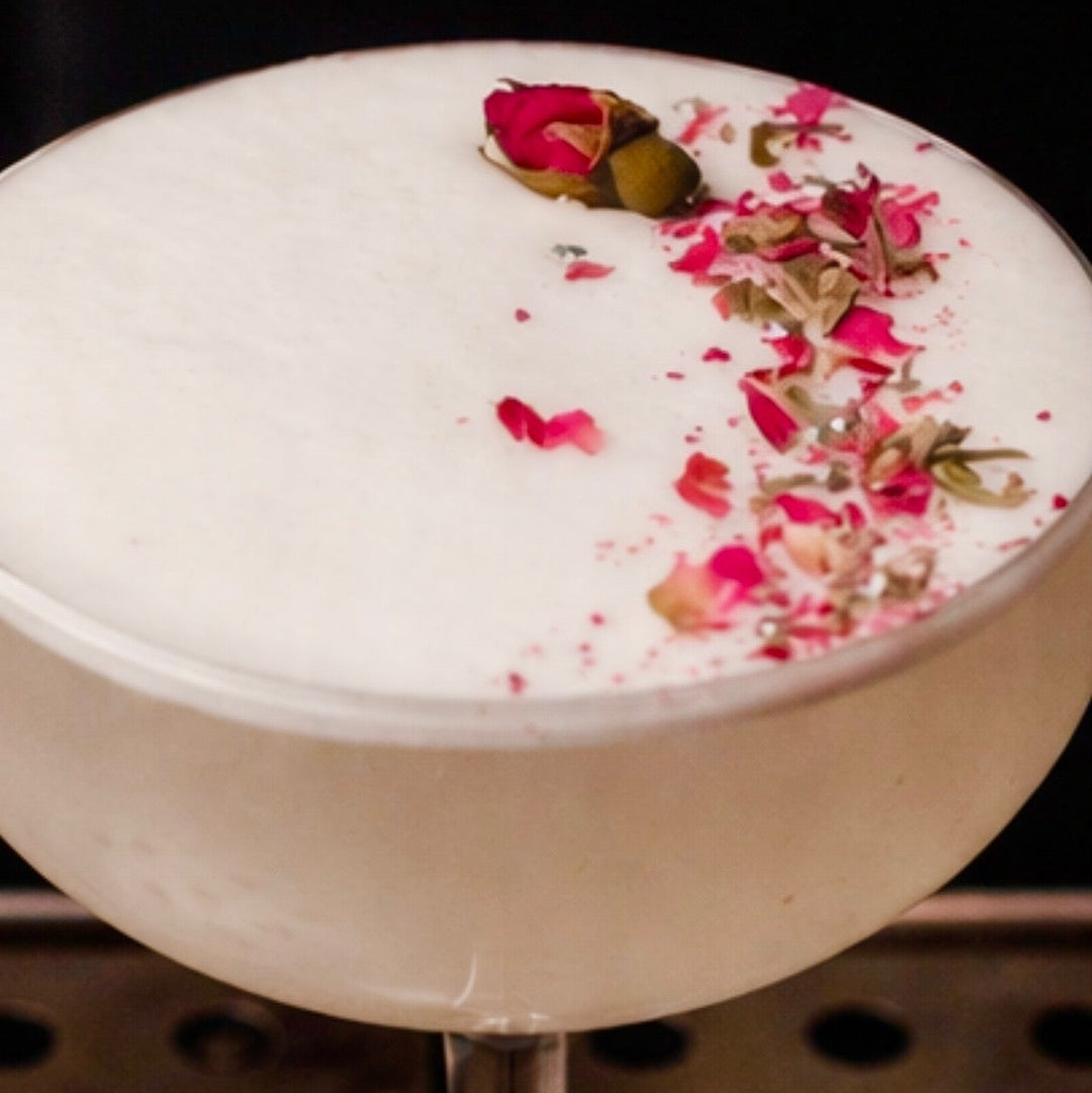 Cocktail with a white foam top and pink floral garnish on a dark background