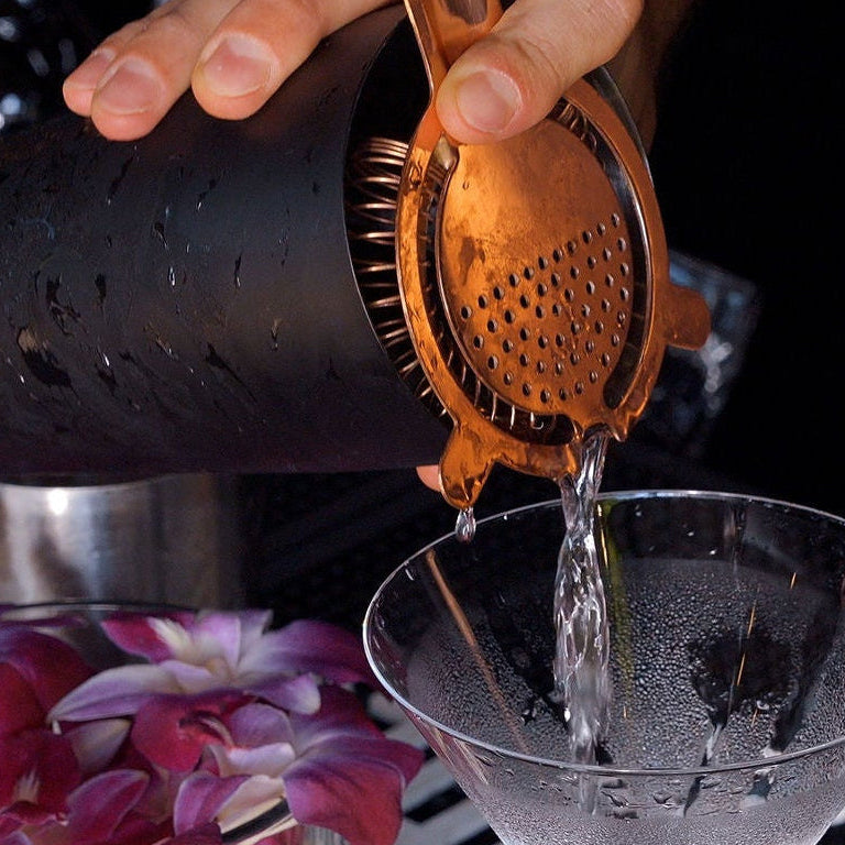 Cocktail being prepared with a shaker and strainer over a glass, surrounded by flowers.