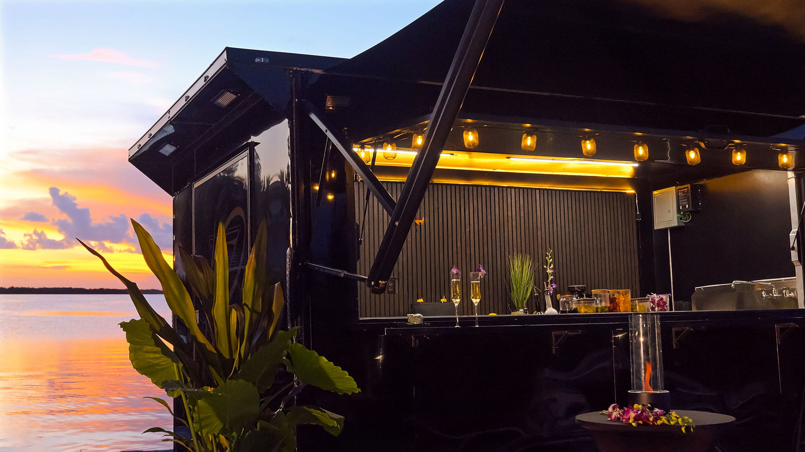 Mobile bar with drinks and decor against a sunset sky and water.