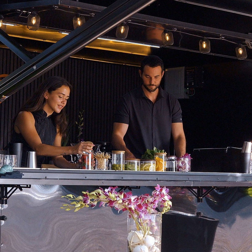 Two bartenders behind a mobile bar counter with floral arrangements and drinks.