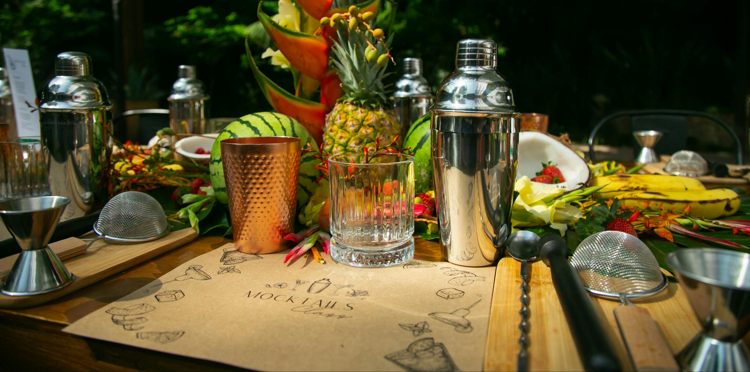 Bar setup with cocktail shakers, glasses, and tropical fruit on a wooden surface outdoors.