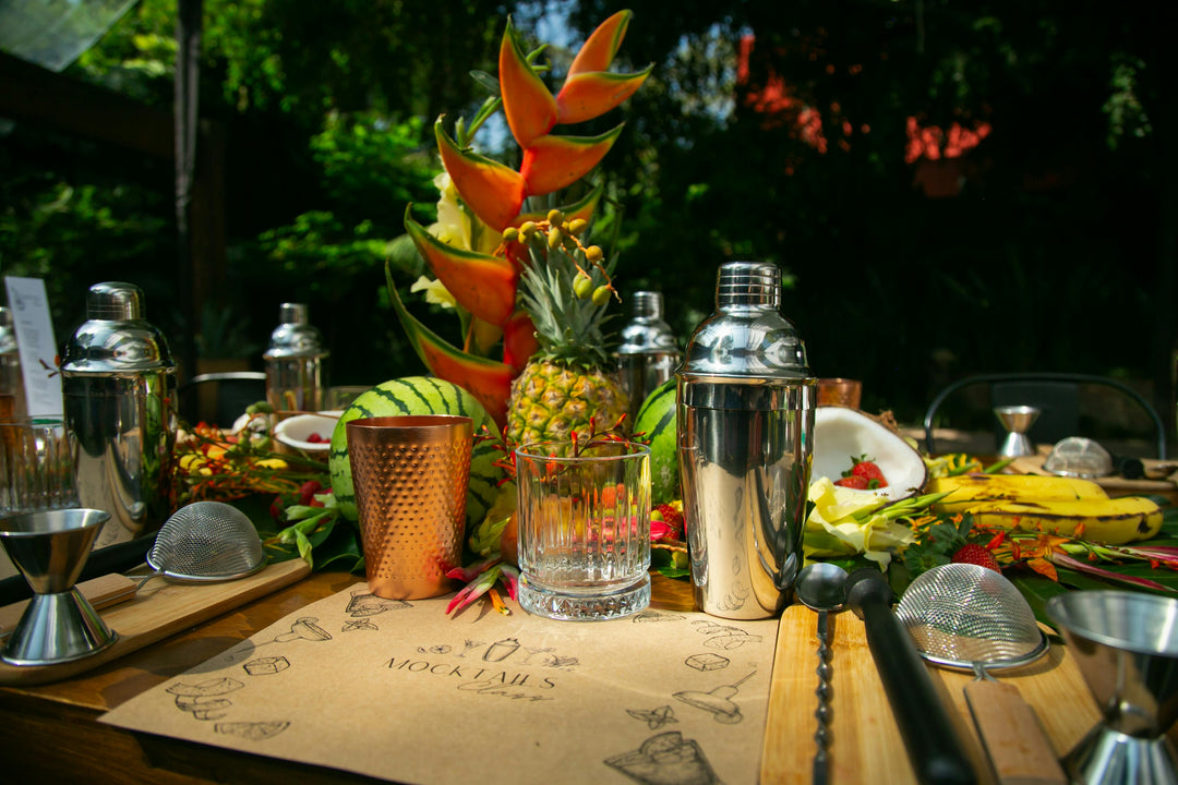 Pop up Bar setup with cocktail shakers, glasses, and tropical fruit on a wooden surface outdoors.