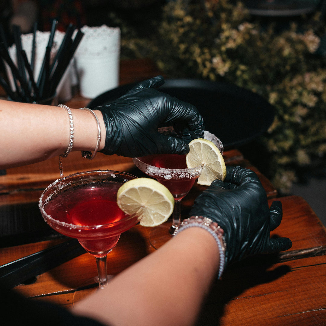 Bartender in black gloves garnishing cocktails with lemon wedges on a wooden bar.